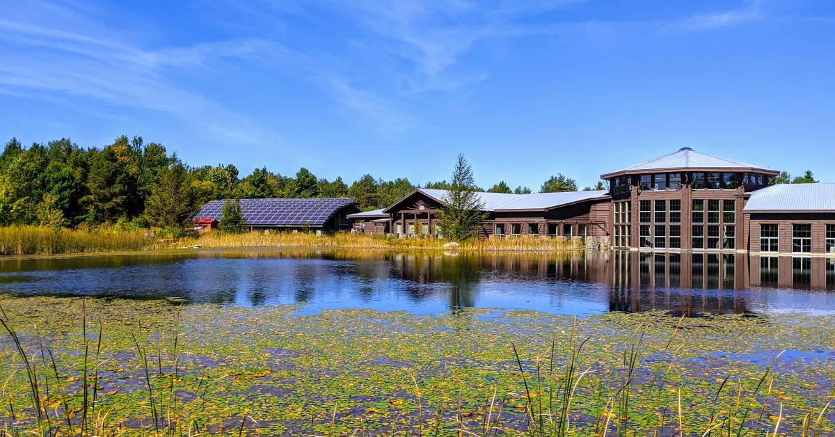 The Wildlife Center in Tupper Lake, featuring a brown building surrounded by greenery, with a serene body of water in the foreground.