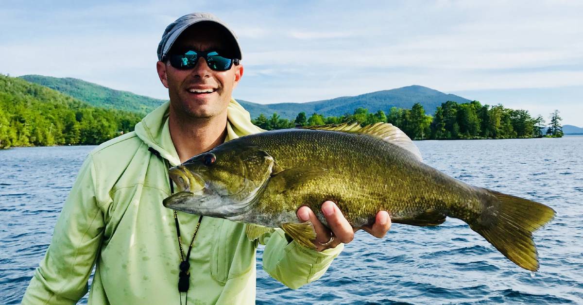 man holds up fish in front of lake george