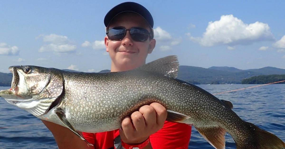 young man holds up large fish in front of lake george