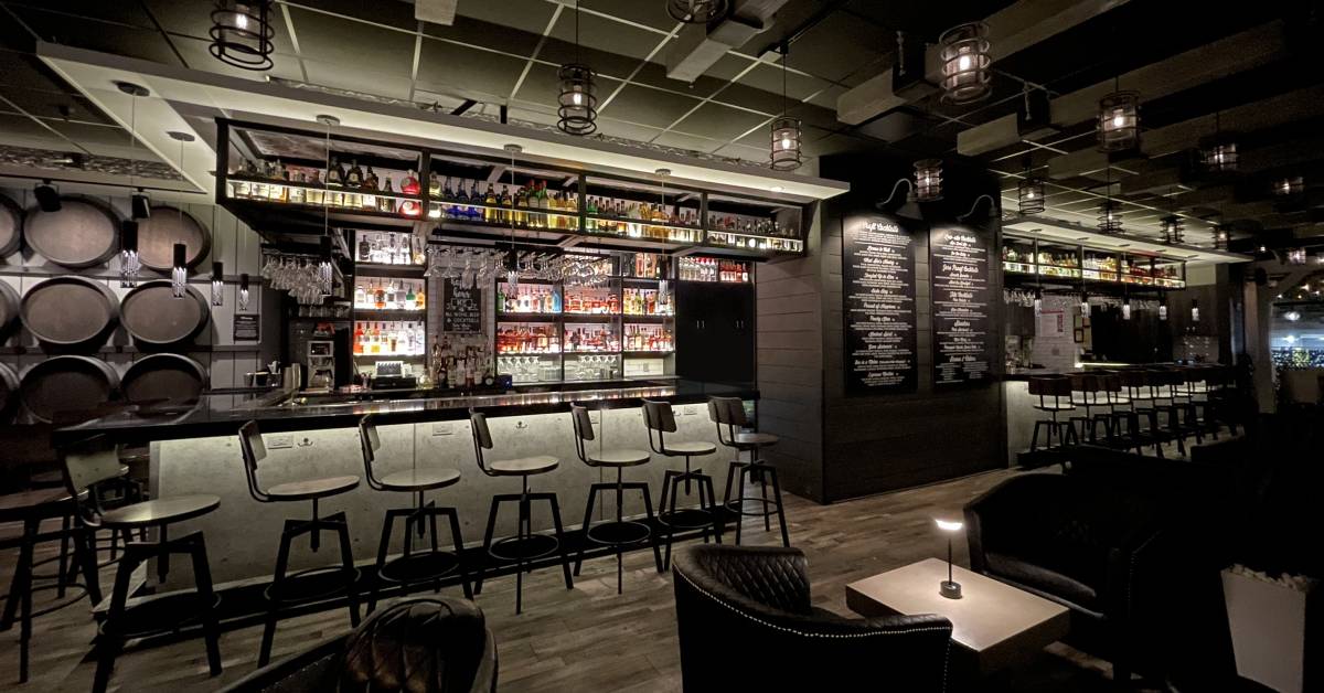 A modern, dimly lit bar with a sleek black countertop, high stools, and illuminated liquor shelves. Cozy leather seating and hanging lanterns create a warm, inviting ambiance.