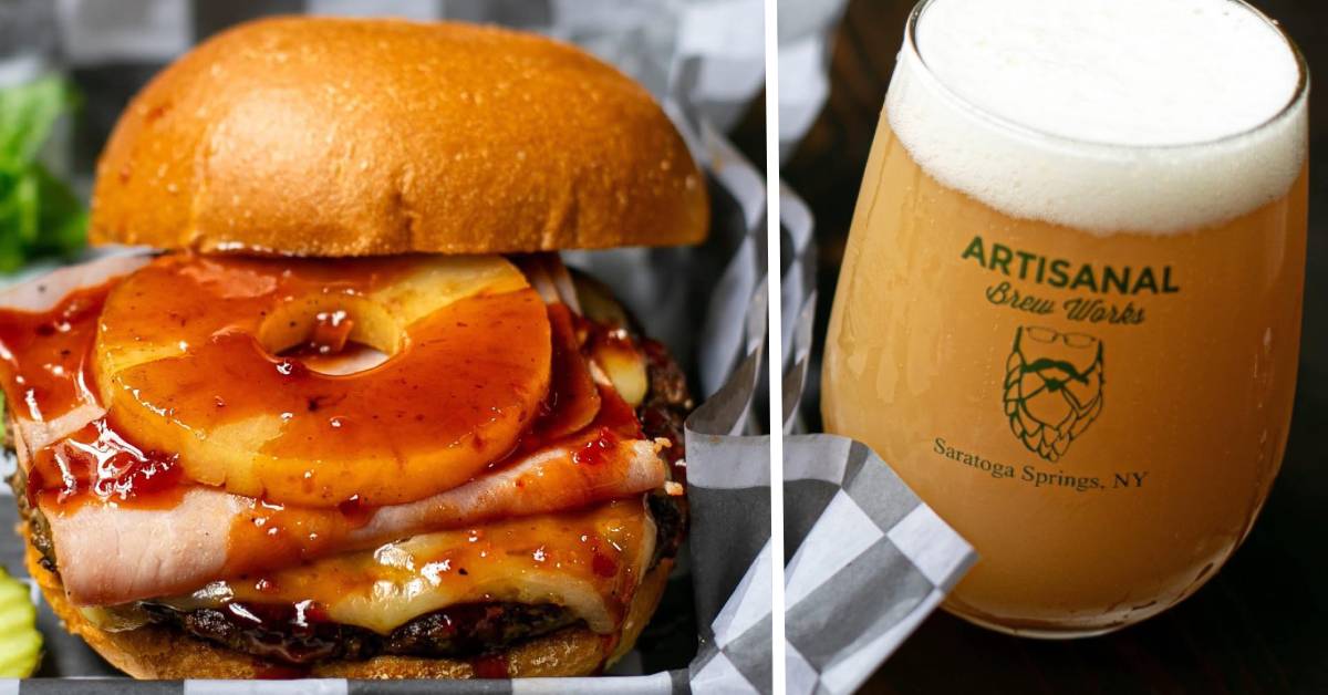 A burger topped with ham, melted cheese, a glazed pineapple ring, and sauce, served on a bun, alongside a hazy craft beer from Artisanal Brew Works in Saratoga Springs, NY.