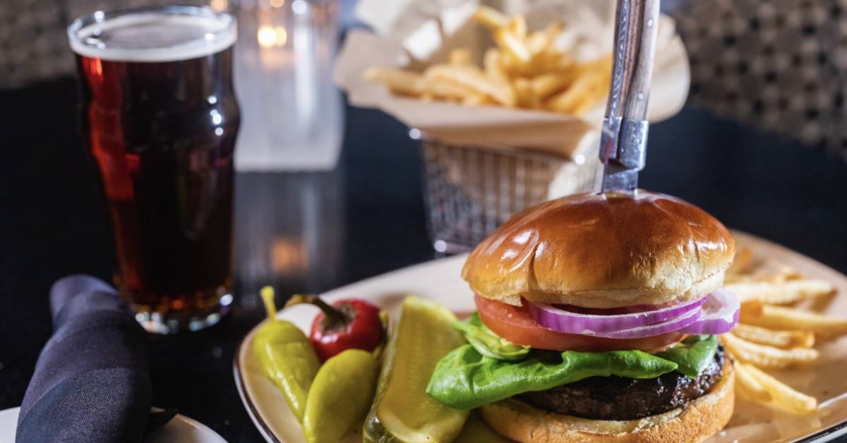 A burger with lettuce, tomato, and onion is served with fries, pickles, and peppers on a plate, accompanied by a dark beer