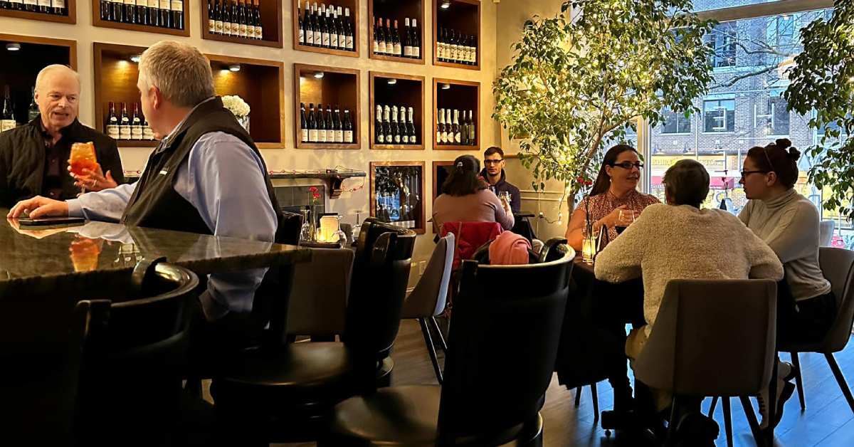 people seated at a wine bar and nearby tables with wines on display on a wall