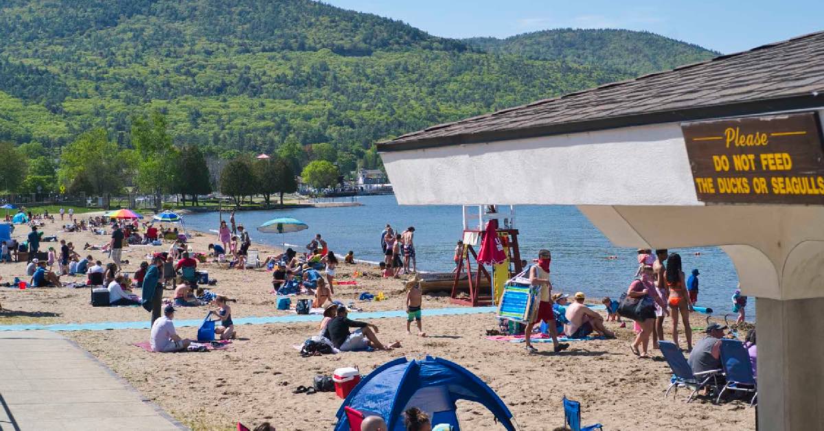 Crowded Million Dollar Beach in Lake George with families on the sand, lifeguard stand, and lake with mountain views behind