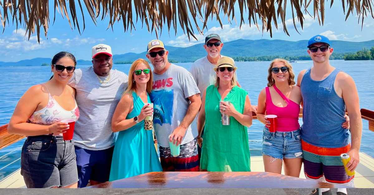 Group of friends enjoying drinks on a tiki tour boat on Lake George, with mountain views and open water in the background