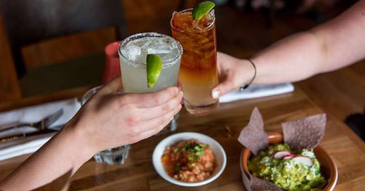 Two people holding drinks with pico, guac, and chips on the table