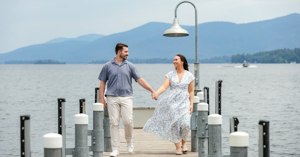 A couple holds hands while walking along a wooden pier with a lake and mountains behind them.