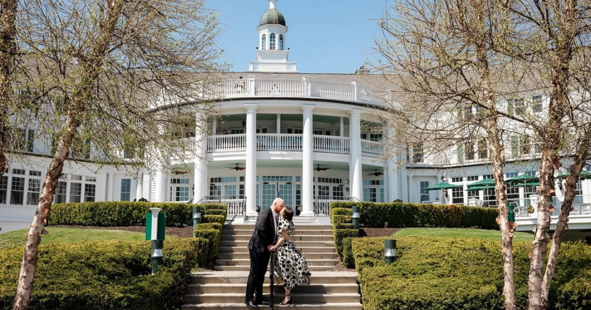 A couple kisses on stone steps leading up to a large white estate featuring a grand columned balcony and cupola.